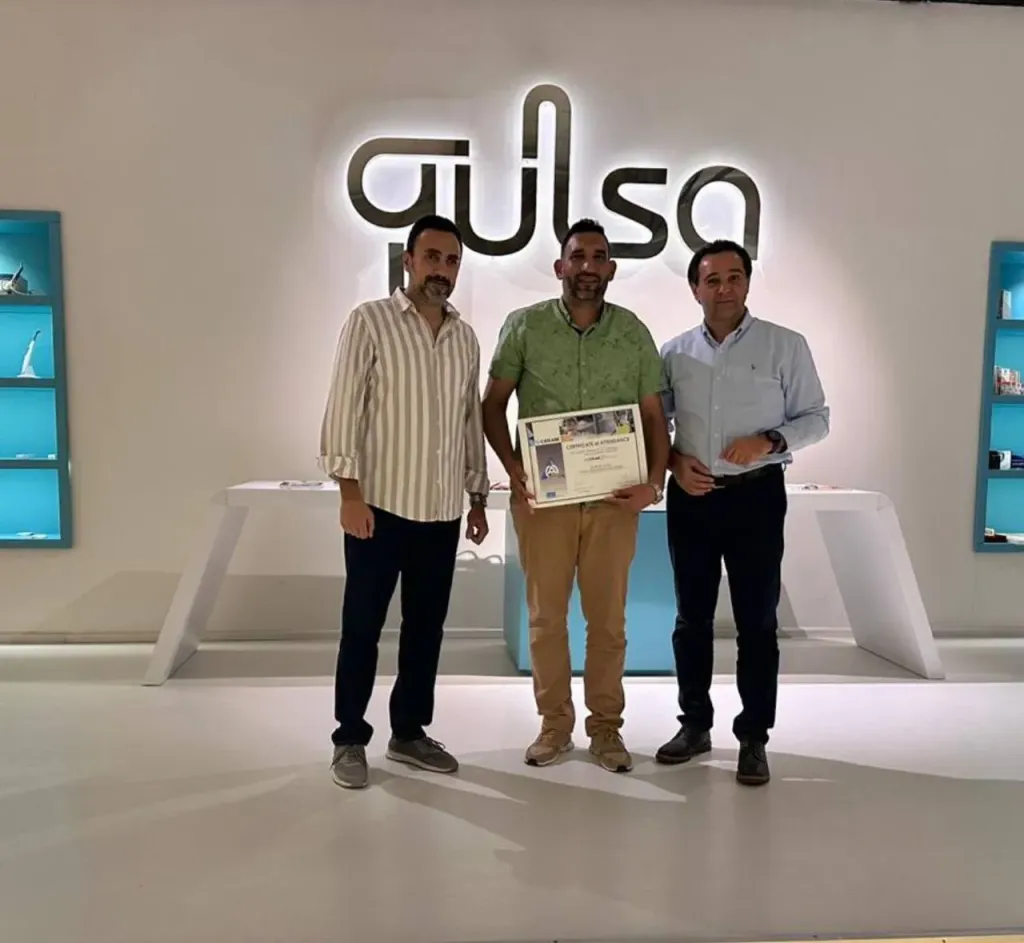 Three men in front of a lit Gulsa sign, the one in the middle holding a certificate of attendance